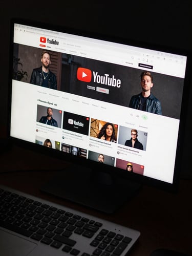 YouTube channel page on a high-resolution monitor showing branded overlay graphics and channel art in a dark edit suite, monitor glow illuminating the keyboard below, overhead angle
