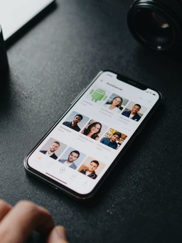 Close-up overhead flat-lay of an Android phone resting on a dark desk, app interface grid visible on screen, natural daylight from a side window casting soft clarity across the surface, hand partially in frame
