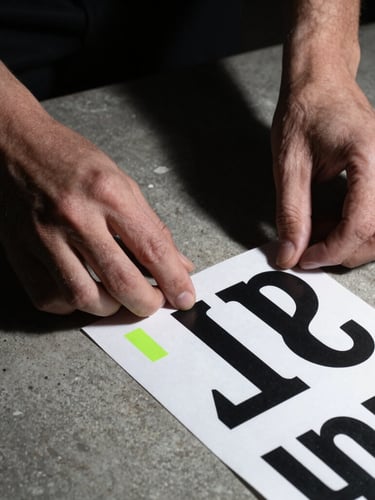 Stark close-up of hands arranging physical typography mockups on a raw concrete surface, harsh direct flash, deep shadows, monochrome, 35mm
