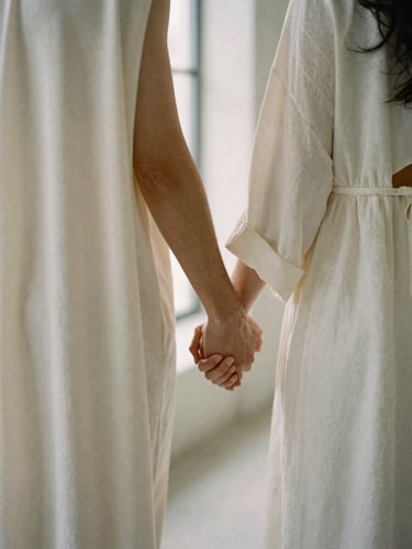 A couple standing hand-in-hand in a minimalist architectural setting, soft window light highlighting the elegant drape of their ivory garments.