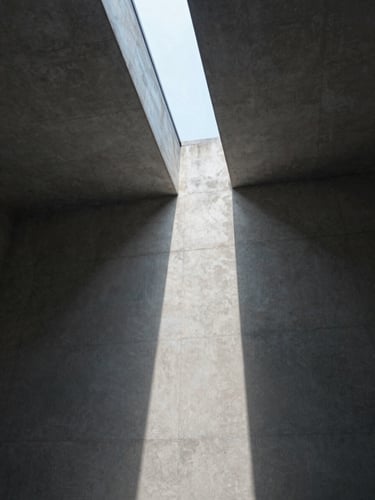 Wide architectural interior shot looking upward at a concrete ceiling where raking natural light enters through a narrow clerestory slot, casting a single sharp line of diffused daylight across the raw surface — no people, no furniture, just the geometry of light and shadow on the structure, photographed in cool north-facing daylight with soft gradation from deep shadow to pale gray stone