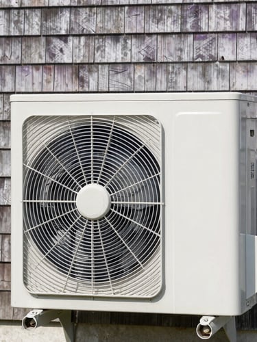 Clean outdoor heat pump coil unit set against weathered cedar shingle siding of a Prince Edward Island cottage, bright natural daylight, crisp focus