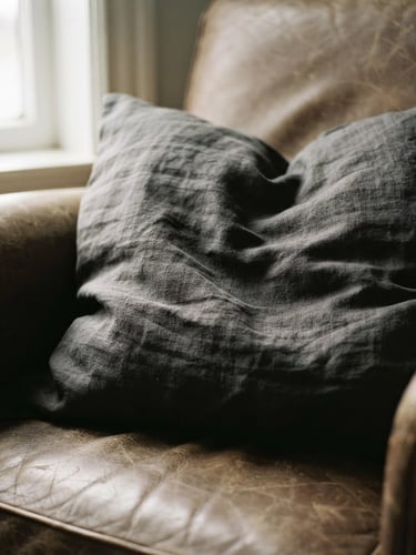 A close-up of a rumpled charcoal linen pillow cover on a worn leather chair, low-angle natural window light casting deep shadows into the heavy fabric grain, warm editorial photography, 35mm lens