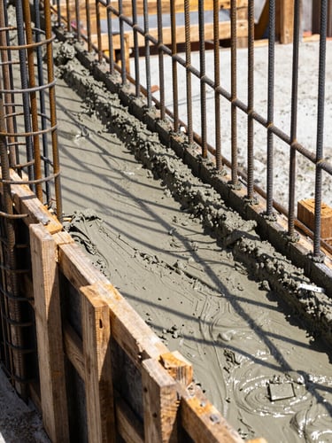 High-contrast close-up photograph of a concrete pour on a real Colombian construction site. Wet concrete flowing into wooden formwork reinforced with steel rebar. Bright natural daylight highlights the texture of the wet cement and the metallic sheen of the steel. Sharp focus, 35mm architectural realism.