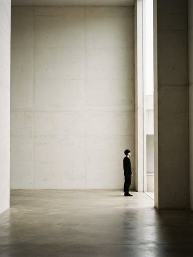 Minimalist editorial photography, soft diffused north-facing daylight. A wide environmental shot of a quiet architectural concrete space with a single contemplative silhouette standing near a tall, narrow opening. Soft shadows, stone gray and muted ivory tones, 35mm film.