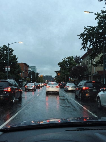Overcast Seattle street view from inside a car, looking through a rain-streaked windshield at the glowing taillights of traffic on a wet evening, cool slate tones, 35mm.