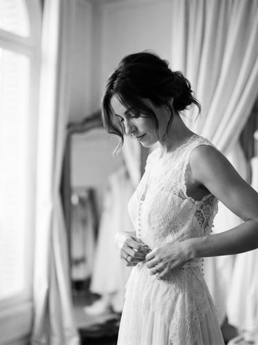 High-contrast black and white editorial portrait of a bride during a fitting, soft natural window light, warm ivory tones, elegant drapery, shallow depth of field.
