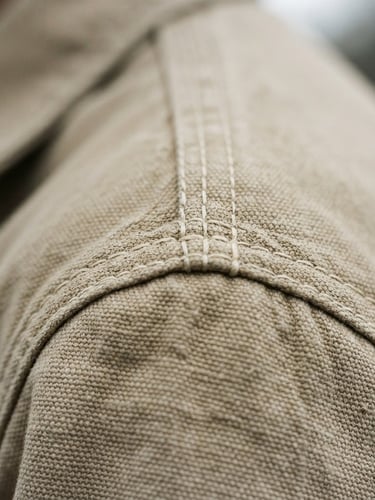 Extreme close-up of heavy-weight cotton stitching on a structured beige shoulder, low-contrast overcast daylight, desaturated neutral tones, 35mm film grain.