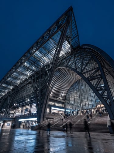 Low-angle heroic shot of a massive steel-and-glass transit hub at blue hour, sharp structural shadows, cool daylight, architectural photography style.