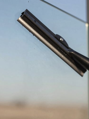 Extreme close-up, tight macro frame: a professional rubber squeegee blade dragging across a large glass pane in bright diffused morning daylight, water beading and sheeting cleanly to one side, glass surface revealing near-perfect clarity with a faint reflection of desert sky, no human face visible, technical and precise