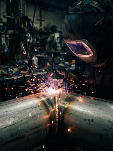 Dramatic close-up of a welder in a dark workshop, bright crimson sparks flying from a clean TIG weld on polished stainless steel, sharp focus, technical lighting