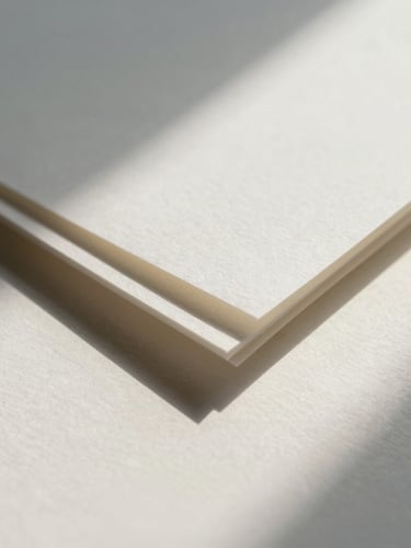 Macro close-up of textured handmade paper with deckled edges, soft architectural shadows casting across a warm white surface, diffused northern daylight.