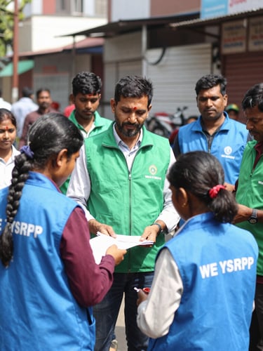 A group of WE YSRCP volunteers in branded vests, actively engaging with Guntur residents during a community outreach event, bright natural daylight, street-level perspective, authentic civic photography, 35mm lens.