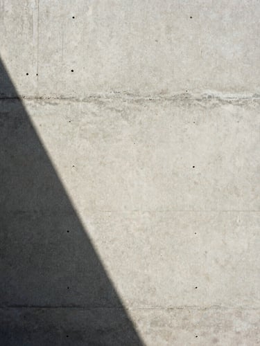 Extreme close-up of raw board-formed concrete wall, sharp diagonal shadow from natural daylight, minimalist editorial framing, medium format film texture