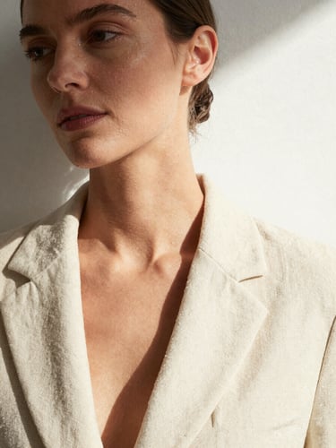 Close-up portrait of a woman in a cream linen blazer against a white studio wall, north-facing daylight raking across her jaw and collarbone, face turned slightly left, negative space on the right third of the frame