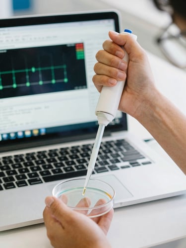 Medium close-up of a student's hands manipulating a pipette over a petri dish, with a laptop screen displaying bioinformatics software in the background, bright natural indoor light, candid, 35mm film style.