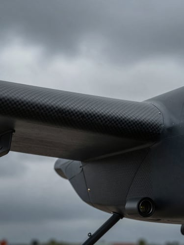 Macro detail shot of a carbon-fiber UAV airframe wing joint and optical sensor payload, matte black finish, cool slate tones, dramatic overcast daylight, 50mm.