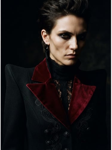 dark editorial fashion photography, close-up of a model wearing a structured sharp-shouldered gothic blazer with deep cherry red velvet lapels, low-key chiaroscuro lighting, dramatic shadows, 35mm