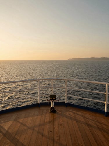 Wide panoramic view from a ship's open deck railing at golden hour, the bow cutting across calm open ocean toward a distant coastline, warm amber light across the deck planking, no people visible — pure environmental scale