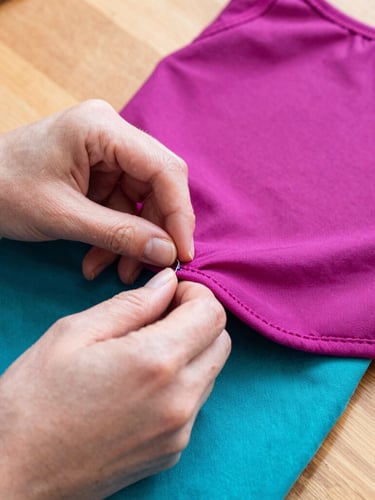 Close-up shot of hands carefully stitching a magenta pet garment on a wooden table, warm daylight, focusing on the neat hidden seams and fabric texture