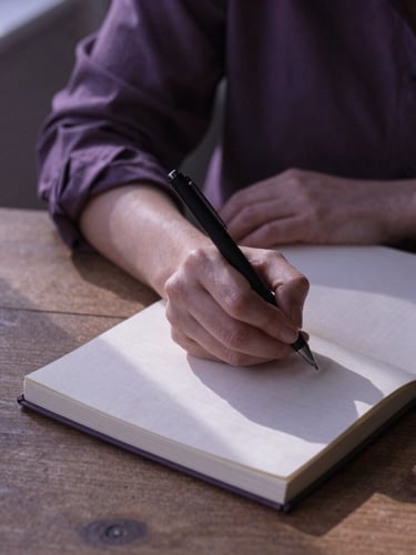 A woman writing in a linen journal on a wooden table, soft shadows on textured paper, diffused natural afternoon light, muted lilac tones, 35mm