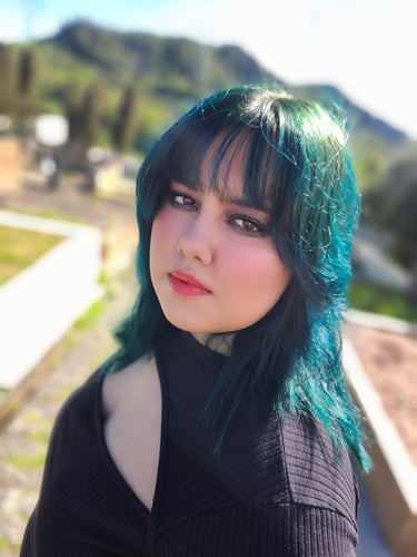 Young woman with blue and green dyed hair and bangs wearing makeup in a sunny outdoor setting.