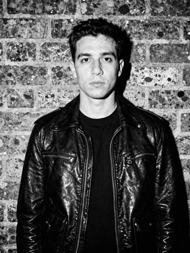 High-contrast monochrome photo of a creator in a leather jacket standing against a gritty brick wall in East London, harsh direct flash, 35mm film grain