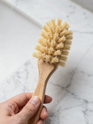 Overhead macro shot of a hand holding a solid beechwood dish brush, natural light reflecting off wet marble tiles, water droplets, crisp editorial photography.