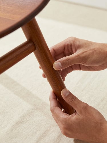 Close overhead view of hands in natural north-facing light carefully inspecting the underside of a mid-century wooden chair leg, examining a maker's mark, sparse cream linen surface beneath, no props