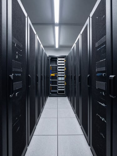 Wide shot from the end of a narrow aisle inside a high-density data center, rows of black server racks receding into the distance under bright overhead LED strips, clinical white light casting hard shadows on cable management panels, tight framing emphasizing depth and density, no people
