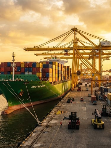 Wide low-angle shot of a green container ship docked at Tanjung Priok port during golden hour, safety yellow cranes towering in the background, high-contrast industrial light, 35mm.