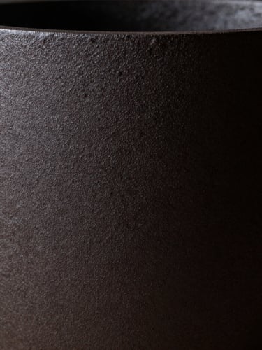 Extreme close-up of a dark, matte cast iron vessel, a single shaft of light revealing its rough, porous texture, deep shadows, minimalist fine art aesthetic.