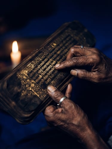 Low-key dramatic photo of hands holding an ancient Sanskrit manuscript under warm candlelight, gold leaf detailing, royal blue shadows, 35mm