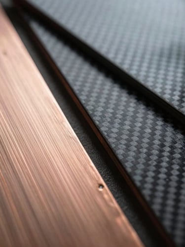 Extreme macro photograph of high-contrast raw material swatches, displaying brushed copper plates and textured carbon fiber sheets, overcast industrial daylight.
