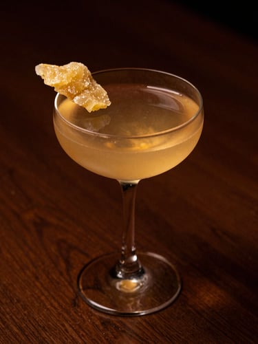 A minimalist cocktail in a delicate coupe glass, garnished with a single slice of crystallized ginger, warm amber candlelight glow, deep shadows