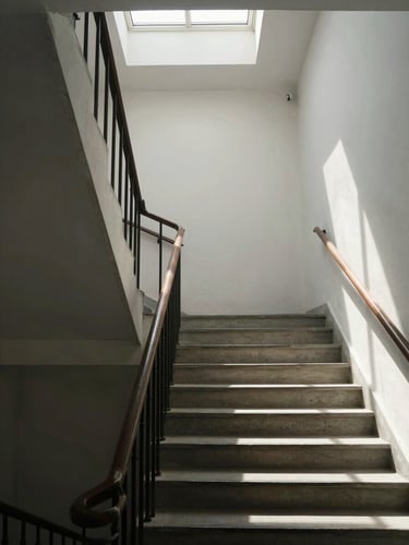 Close environmental shot looking up a clean concrete staircase inside a Klaipėda residential building, natural light filtering in from a skylight above, geometric shadow lines on pale walls, no people present