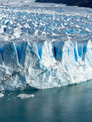 Tall portrait environmental frame—Perito Moreno glacier wall rising from deep aquamarine water, late-afternoon side light raking across the ice face, revealing blue fissures and melt channels, vast and still, no people