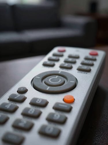 Crisp macro photography of a physical TV remote control, focused on the power button and orange indicator LED, moody living room ambient light, shallow depth of field, 50mm.