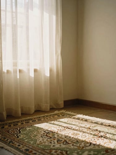 A quiet, sunlit corner of a residential room with soft woven textiles, a prayer rug, and warm light streaming through a sheer curtain.