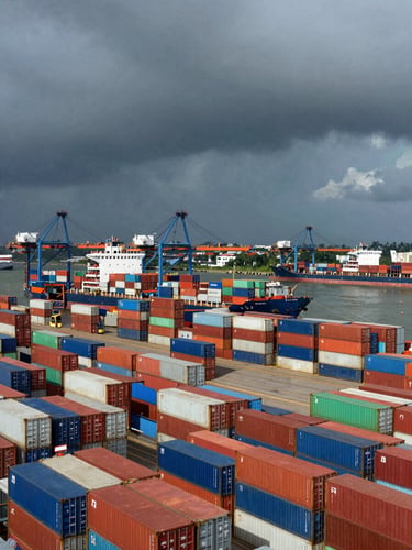 Wide panoramic view of Cochin port under heavy monsoon skies, massive shipping containers stacked in neat grids, container vessel docked, high-contrast natural light with deep shadows, no people.