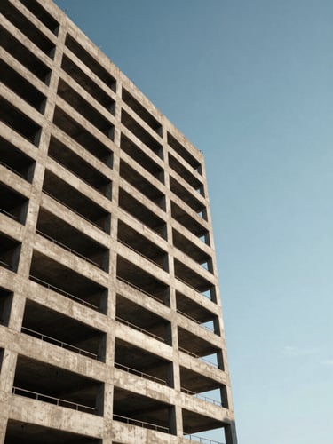 Wide panoramic frame, left-anchored: completed mid-rise commercial building exterior shot from ground level looking upward, concrete facade with exposed structural grid, natural afternoon sidelight casting sharp shadows across the surface, no people, no signage, generous sky in upper right third