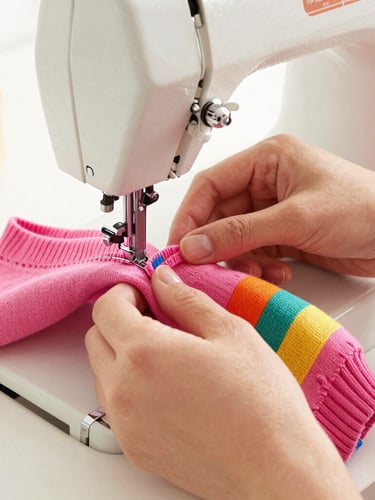 A warm, bright close-up of a tailor's hands carefully stitching a colorful pink pet sweater on a vintage sewing machine, soft focus, warm natural daylight.