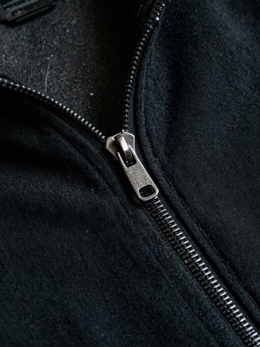 Dramatic low-key close-up of heavy-gauge black cotton fabric textures, a polished metal zipper detail, dark concrete background, sharp focus, 35mm film grain