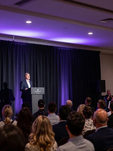 Panoramic corporate event mid-action — a speaker at a podium under dramatic stage lighting, purple-washed overhead wash, audience in soft focus in the foreground, wide enough to show the venue scale