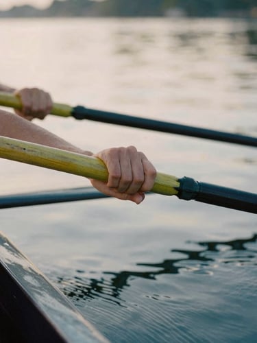 A close-up shot of hands gripping wooden oars in perfect synchronization, soft morning daylight reflecting off calm water, warm editorial photography, muted navy and golden tones, shallow depth of field, 35mm film.