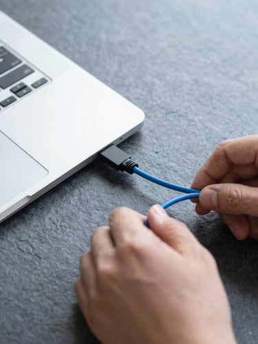 Overhead shot of a clean technical workspace with hands carefully connecting a blue cable to a disassembled laptop, cool daylight, slate background.