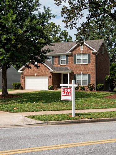 Wide shot of a well-kept suburban street in Lake Wylie SC at midday, a for-sale sign in the front yard of a two-story brick home with a two-car garage, mature trees lining the sidewalk, natural daylight, documentary framing