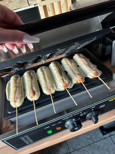 Six golden corn dogs cooking on a commercial waffle hot dog maker machine on a kitchen counter.