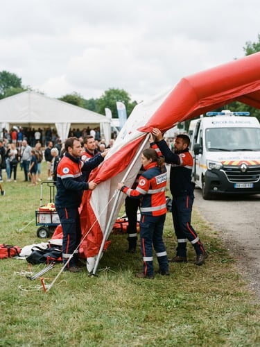 Équipe de secouristes de la Croix Blanche de Loon-Plage en uniforme, déployant une tente de secours sur un événement en extérieur, avec un véhicule d'intervention en arrière-plan, sous un ciel couvert naturel.