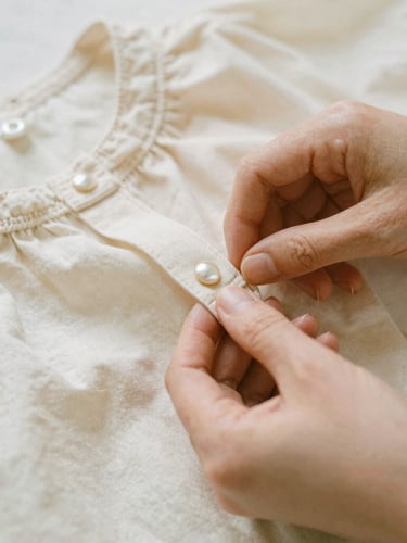 Close-up of hands sewing a delicate collar on a soft ivory linen child's blouse, mother-of-pearl buttons visible, warm diffused natural light, shallow depth of field.